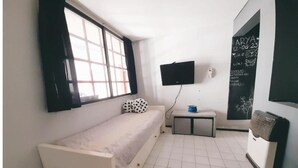Una habitación pequeña con cama individual, ventana, televisor de pantalla plana, una mesita y una pizarra con anotaciones.