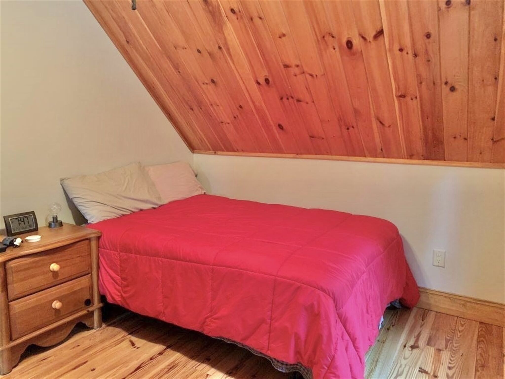 Un dormitorio con techo inclinado de madera, una cama individual con colcha roja, una mesita de noche con un reloj y un aparador de madera.