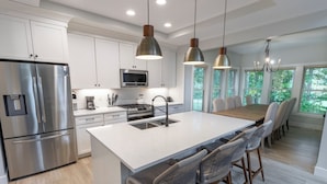 A modern kitchen with white cabinets, stainless steel appliances, a large island with bar stools, pendant lighting, and large windows offering a view of greenery. - Massive New Home w/Community Pool, Game Room (New Buffalo)
