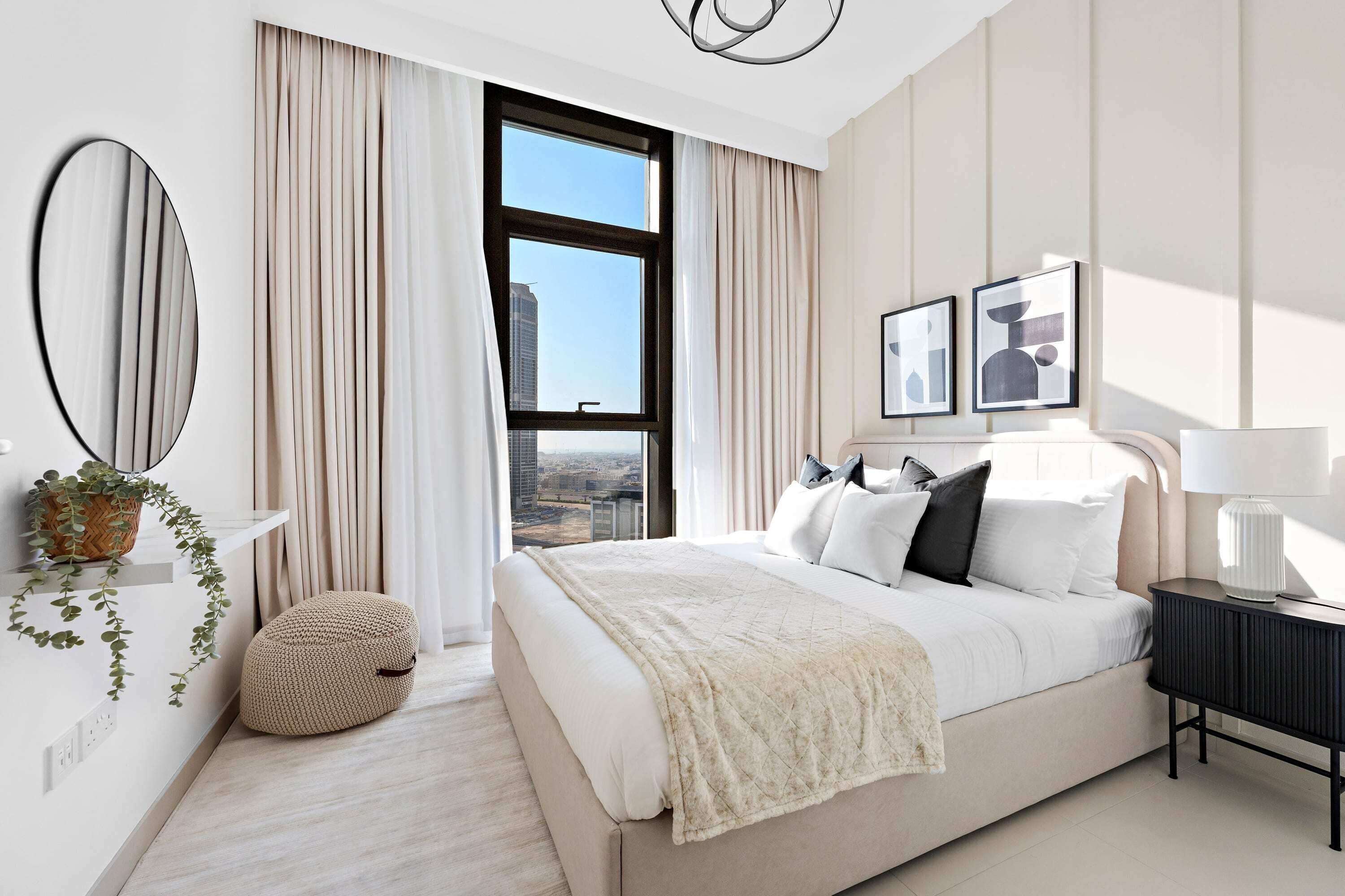 A modern hotel room with a large bed, a bedside table, a mirror, and a view of the city through a large window.