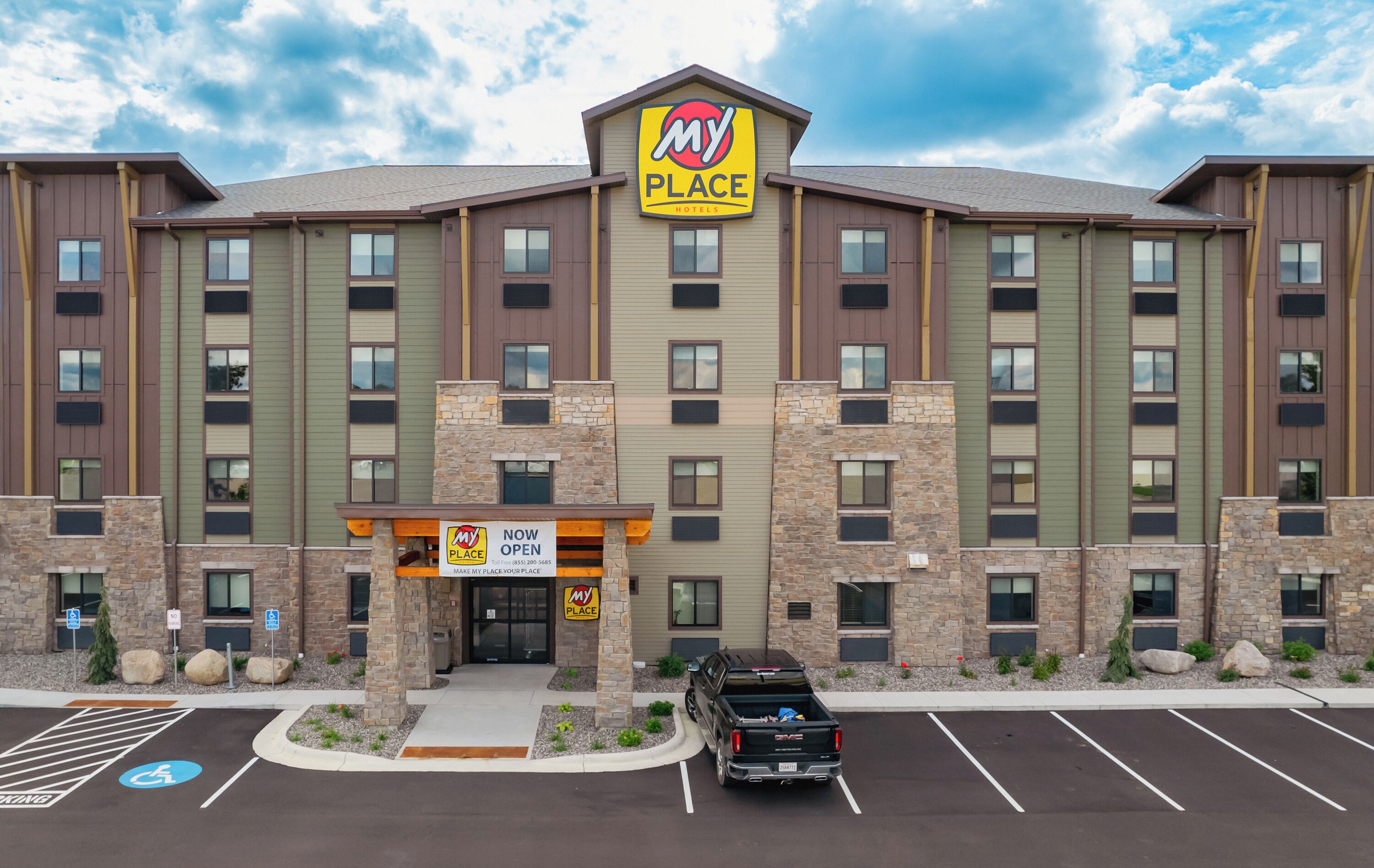 A multi-story hotel with a 'My Place' sign, a parking lot, and a 'Now Open' sign.