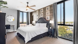 A hotel room with a large bed, a ceiling fan, a desk, a chair, a nightstand with a lamp, and a view of the city through large windows.
