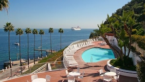 Ein Poolbereich mit einem gewundenen Pool, weißen Liegestühlen und Blick auf das Meer mit Booten.