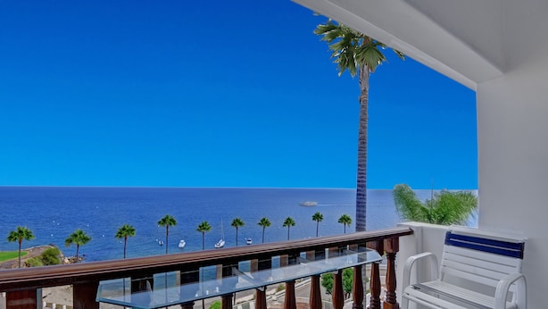 Ein Balkon mit Glasgeländer blickt auf einen Strand mit Palmen und Booten.