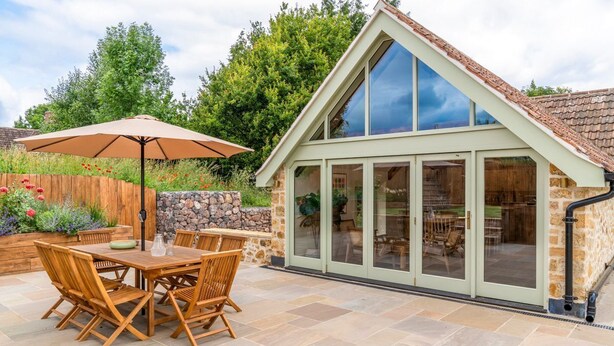 A patio area with a wooden table and chairs, a large umbrella, and a stone wall. - Little Hideaway - Four Bedroom Villa, Sleeps 8 (Whitelackington)