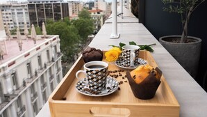 Een dienblad met een koffiekop, muffin en een roos op een balkon met uitzicht op een stadslandschap.