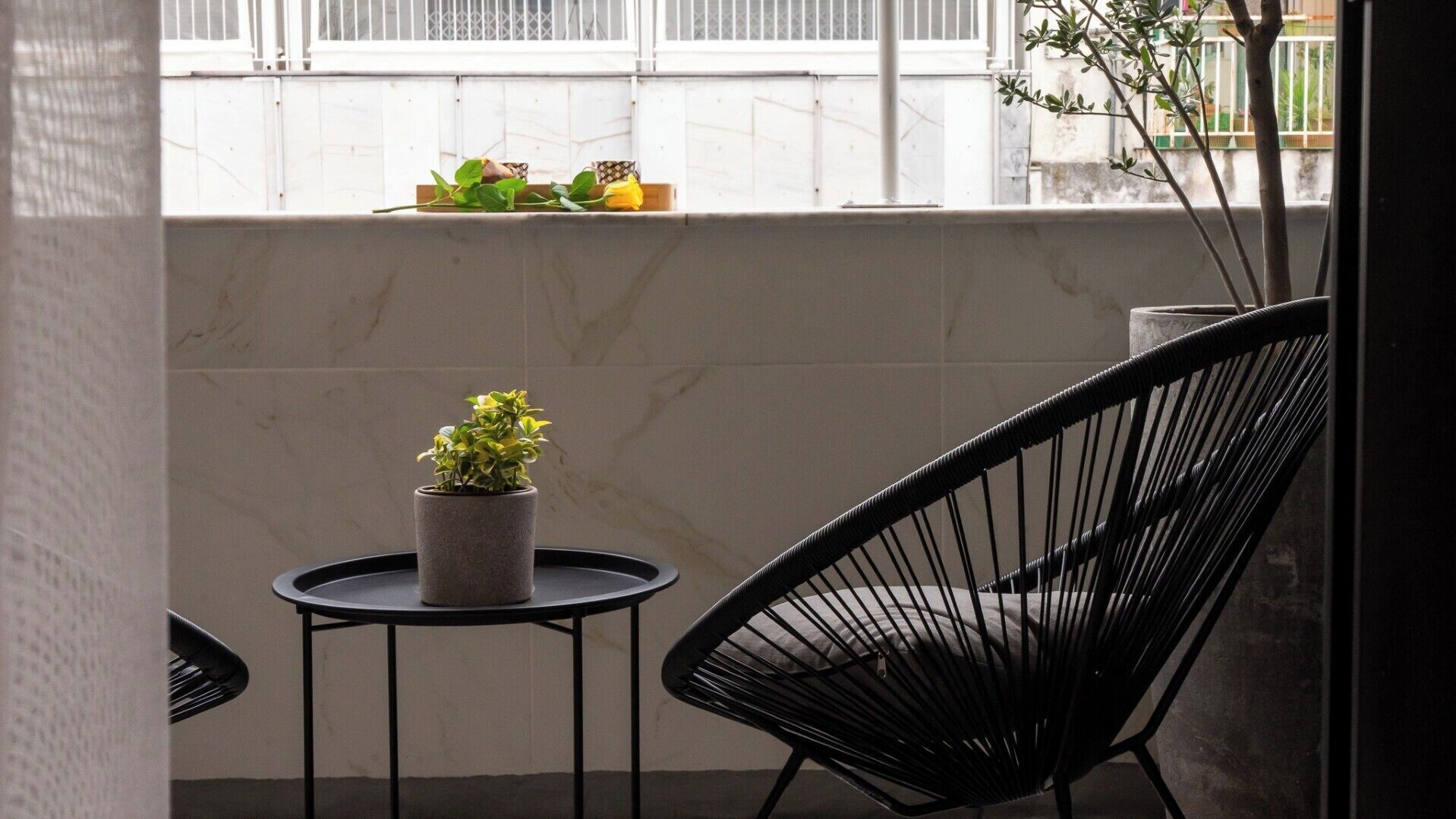 A small balcony with a round table, a potted plant, a black chair, and a window with a view of a building and a tree.