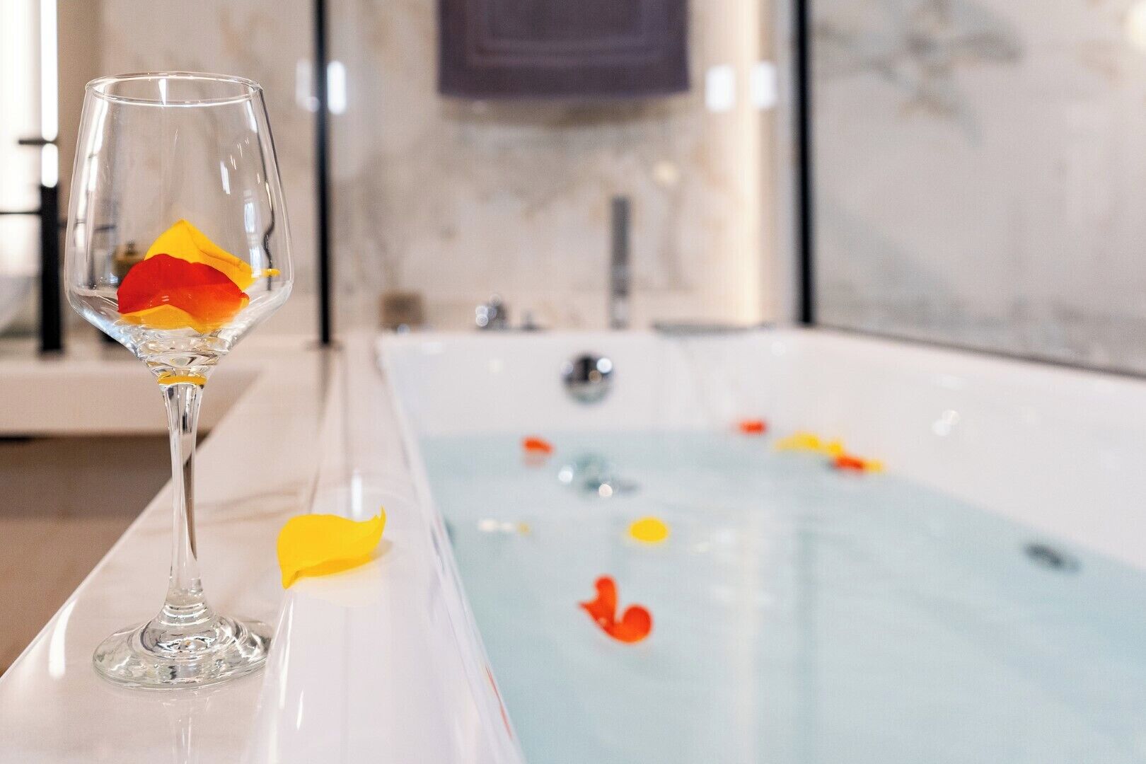 A clear glass with a rose and a yellow flower on a white surface, a bathtub with water and rose petals, and a marble wall in the background.