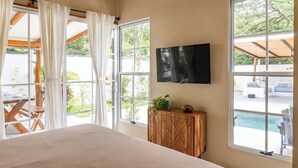 Ein Schlafzimmer mit einem großen Bett, einem an der Wand befestigten Fernseher und Blick auf einen Außenpool durch große Fenster.