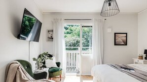 A bedroom with a bed, a TV mounted on the wall, a chair, a plant, and a balcony with curtains. - Pensionat Enehall (Bastad)