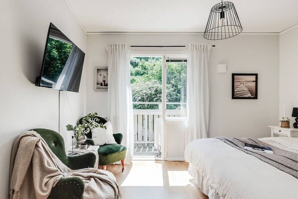 A bedroom with a bed, a TV mounted on the wall, a chair, a plant, and a balcony with curtains. - Pensionat Enehall (Bastad)