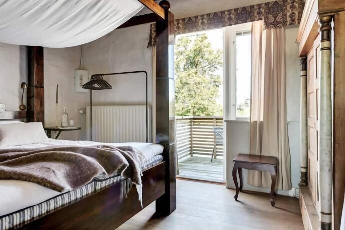 Ein Schlafzimmer mit einem Himmelbett, einem Nachttisch, einer Lampe und Blick ins Freie durch ein Fenster.