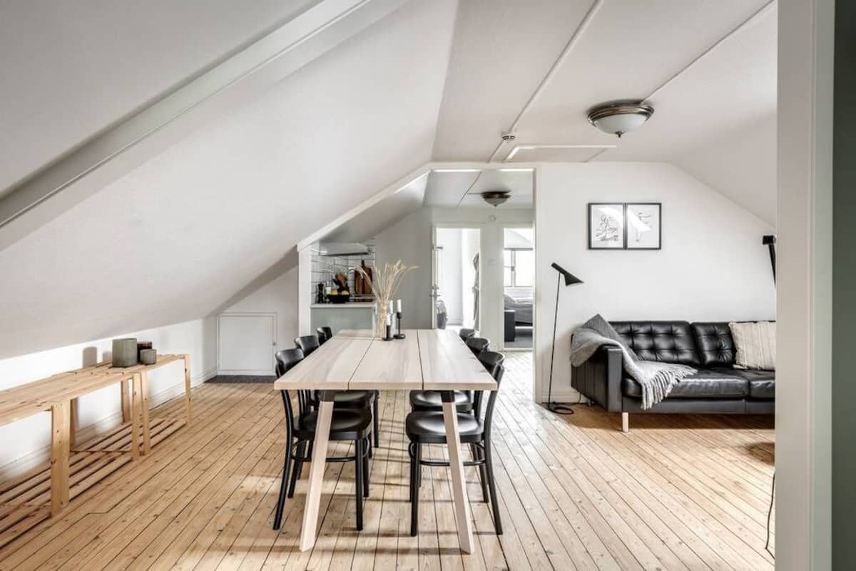 Ein geräumiges Loft mit hoher Decke, einem Essbereich mit langem Tisch und Stühlen, einem schwarzen Ledersofa und einem Holzboden.