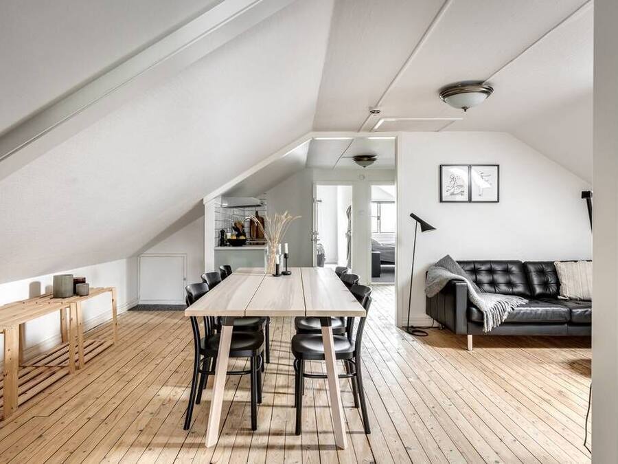 A spacious loft with a high ceiling, a dining area with a long table and chairs, a black leather sofa, and a wooden floor.