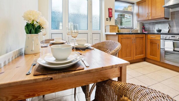 A dining area with a wooden table set for two, a wicker chair, and a view of the kitchen with wooden cabinets and a stainless steel range hood.