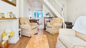 A spacious living area with two beige armchairs, a wooden coffee table, and a white sofa. A staircase leads to an upper level, and a bookshelf is visible in the background.