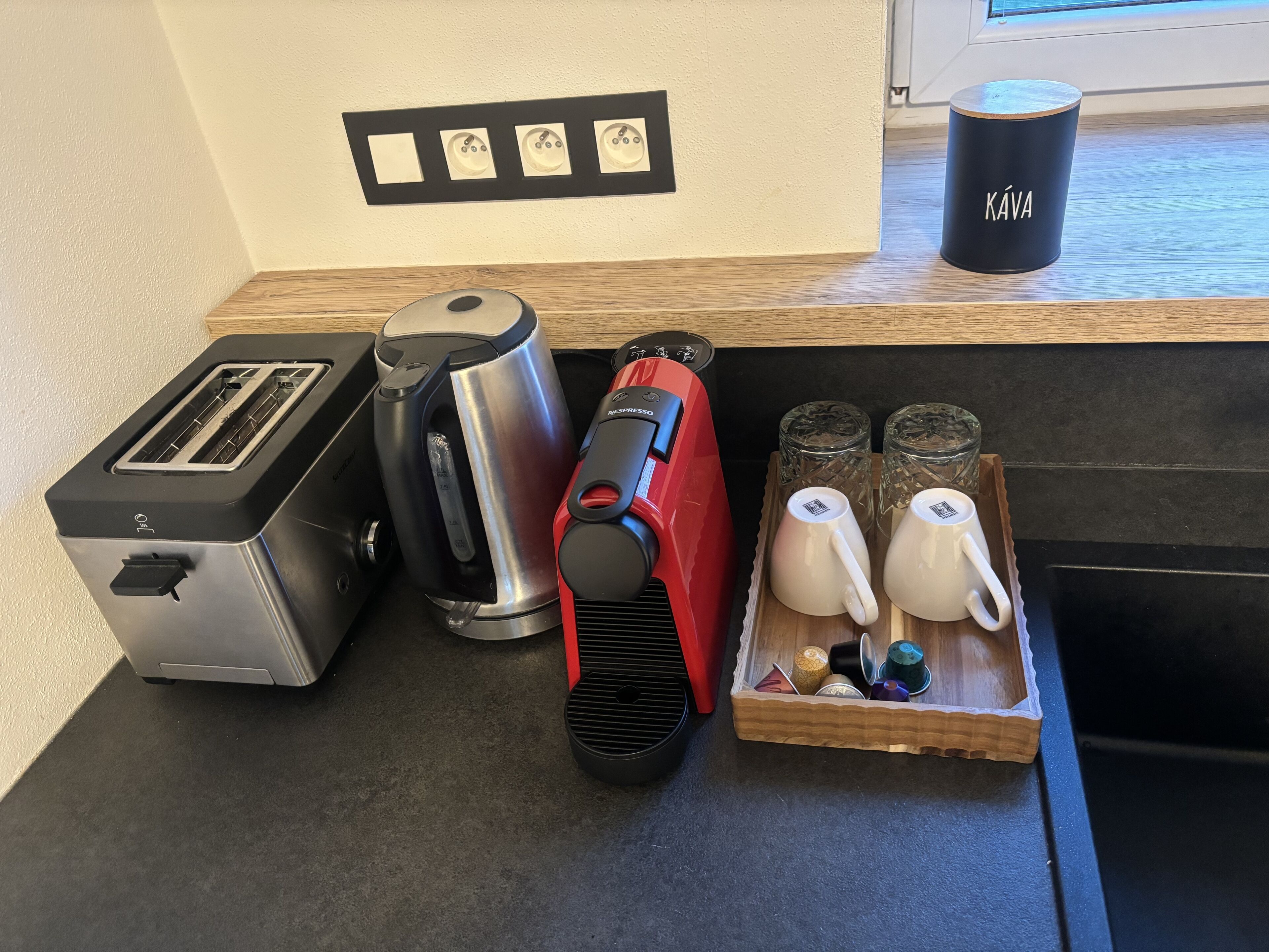 Un plan de travail de cuisine avec un grille-pain, une bouilloire, une cafetière rouge et un plateau contenant des théières et des tasses.