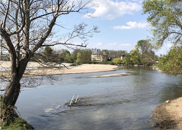 Un río con orilla de arena y un árbol en primer plano. Al fondo, se observa un edificio con una torre.