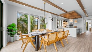Un comedor amplio con una larga mesa y sillas de madera, ventanas grandes con vistas a la vegetación, y una cocina moderna al fondo.