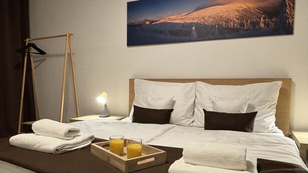 Una cama bien tendida con ropa de cama blanca y marrón, una bandeja con dos vasos de jugo y una escalera de madera apoyada en la pared.