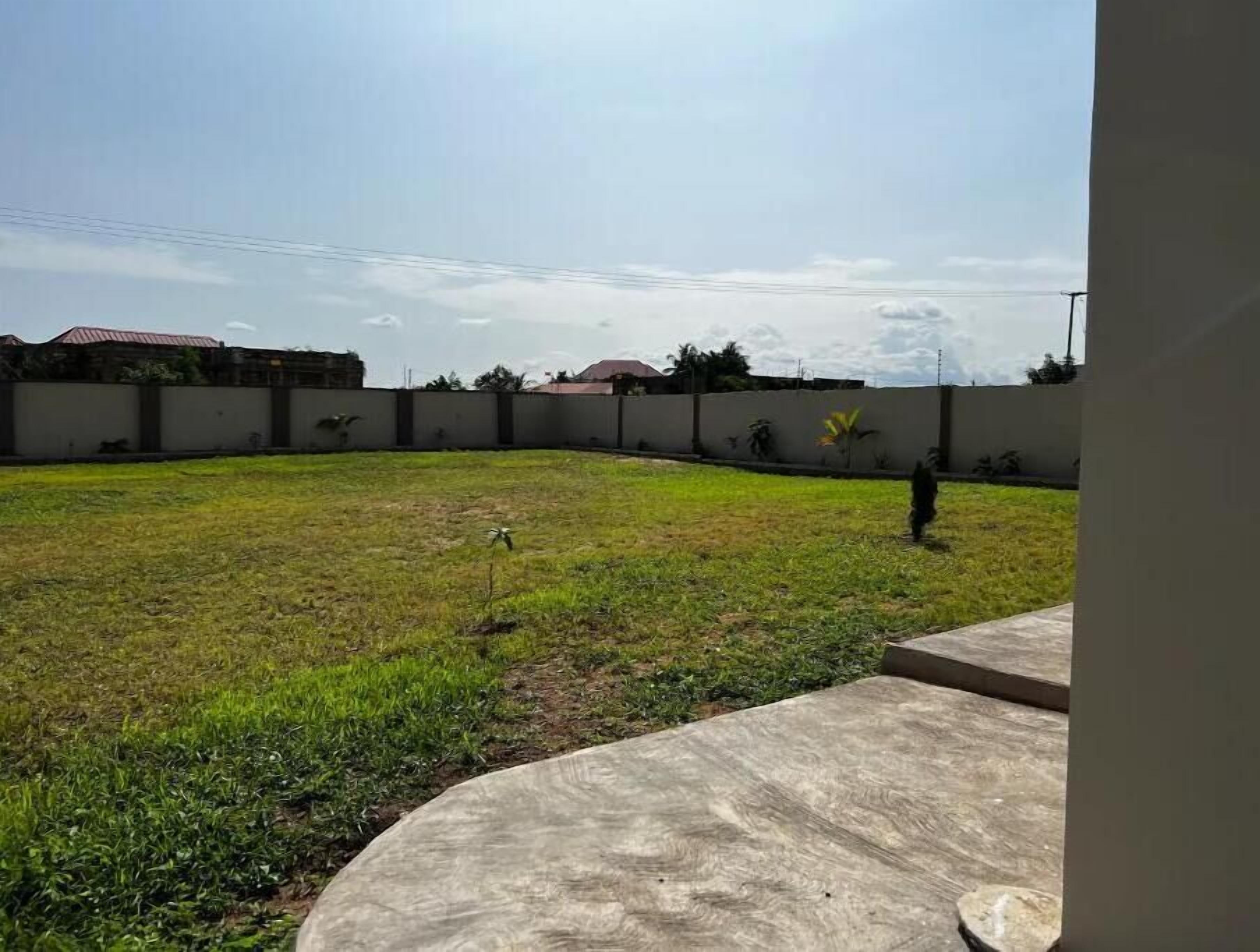 A grassy outdoor area with a concrete patio, surrounded by a low wall and a few trees.