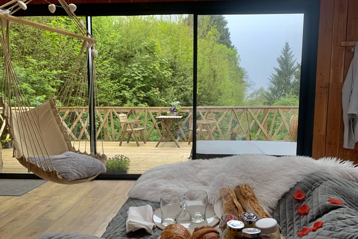 A bedroom with a large window, a hanging chair, a bed with a fluffy blanket, and breakfast items on the bed.