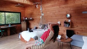 A wooden cabin interior with a hammock bed, a dining area, and a view through a large window. - DOMAINE RÊVES ETOILÉS (La Roche-Maurice)