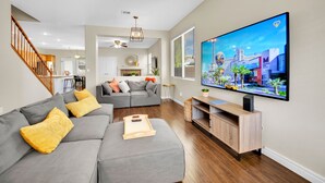 A modern living room with a large flat-screen TV, grey sofas, wooden flooring, and a staircase. - Seven Palms Stylish Home with Pool, Hot Tub,& Movie Room (Henderson)