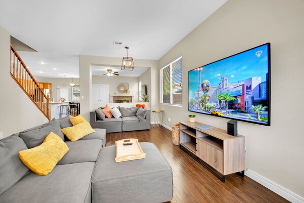 A modern living room with a large flat-screen TV, grey sofas, wooden flooring, and a staircase. - Seven Palms Stylish Home with Pool, Hot Tub,& Movie Room (Henderson)