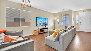 A modern living room with a large flat-screen TV, grey sofas, wooden flooring, and a ceiling light fixture. - Seven Palms Stylish Home with Pool, Hot Tub,& Movie Room (Henderson)