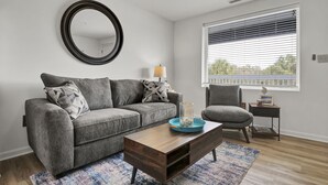 Un salon moderne avec un canapé gris, une table basse en bois, un tapis bleu, une table d’appoint avec une lampe et un fauteuil.