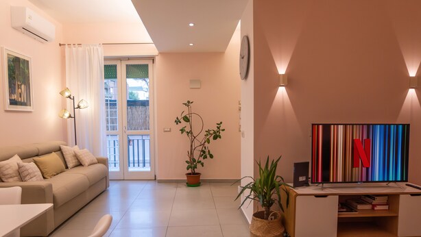 A modern living room with a sofa, a dining table, and a TV. There is a potted plant and a wall-mounted clock. - La Casupola - Gemelli Garden

 (Roma)