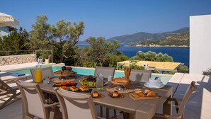 Une table à manger dressée avec de la nourriture et des boissons, donnant sur une piscine et la mer.