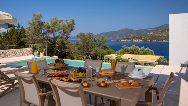 Mesa de comedor dispuesta con comida y bebidas con vistas a una piscina y al mar.