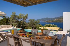 Ein Esstisch gedeckt mit Speisen und Getränken mit Blick auf einen Pool und das Meer.