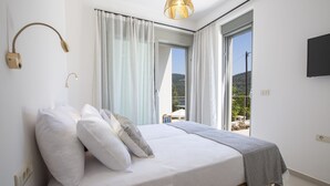 Un dormitorio con cama, ropa de cama blanca, una lámpara de noche, un televisor y vistas al exterior.