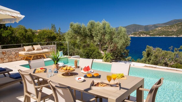 Un área de comedor junto a la piscina con una mesa preparada para dos, con vista al mar azul y un paisaje montañoso.