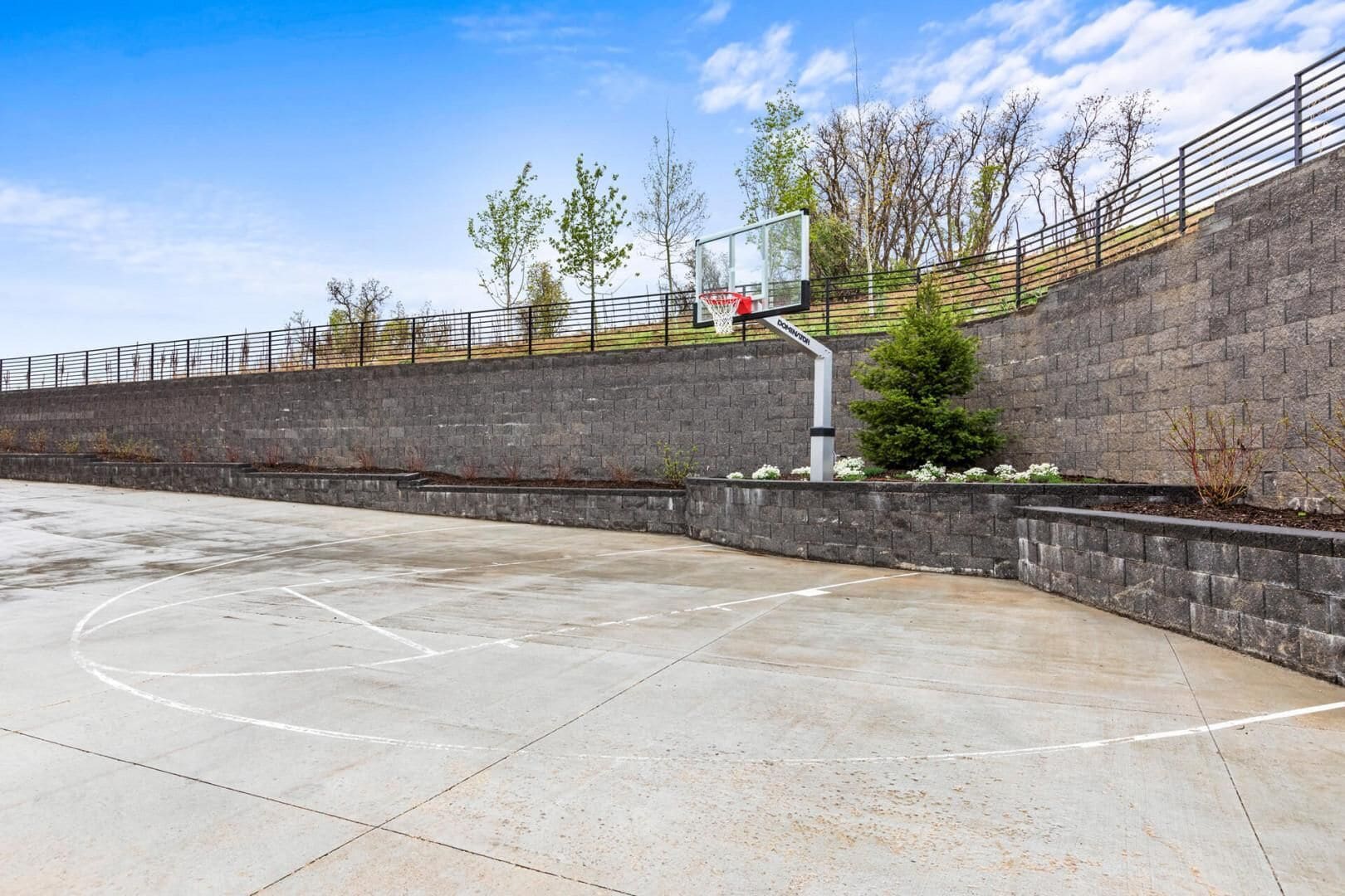 An outdoor basketball court with a hoop, surrounded by a stone wall and a metal fence.