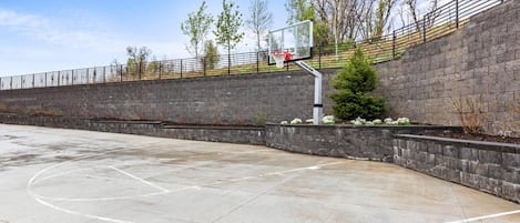Un terrain de basket en plein air, doté d’un panier, entouré d’un mur en pierre et d’une clôture métallique.