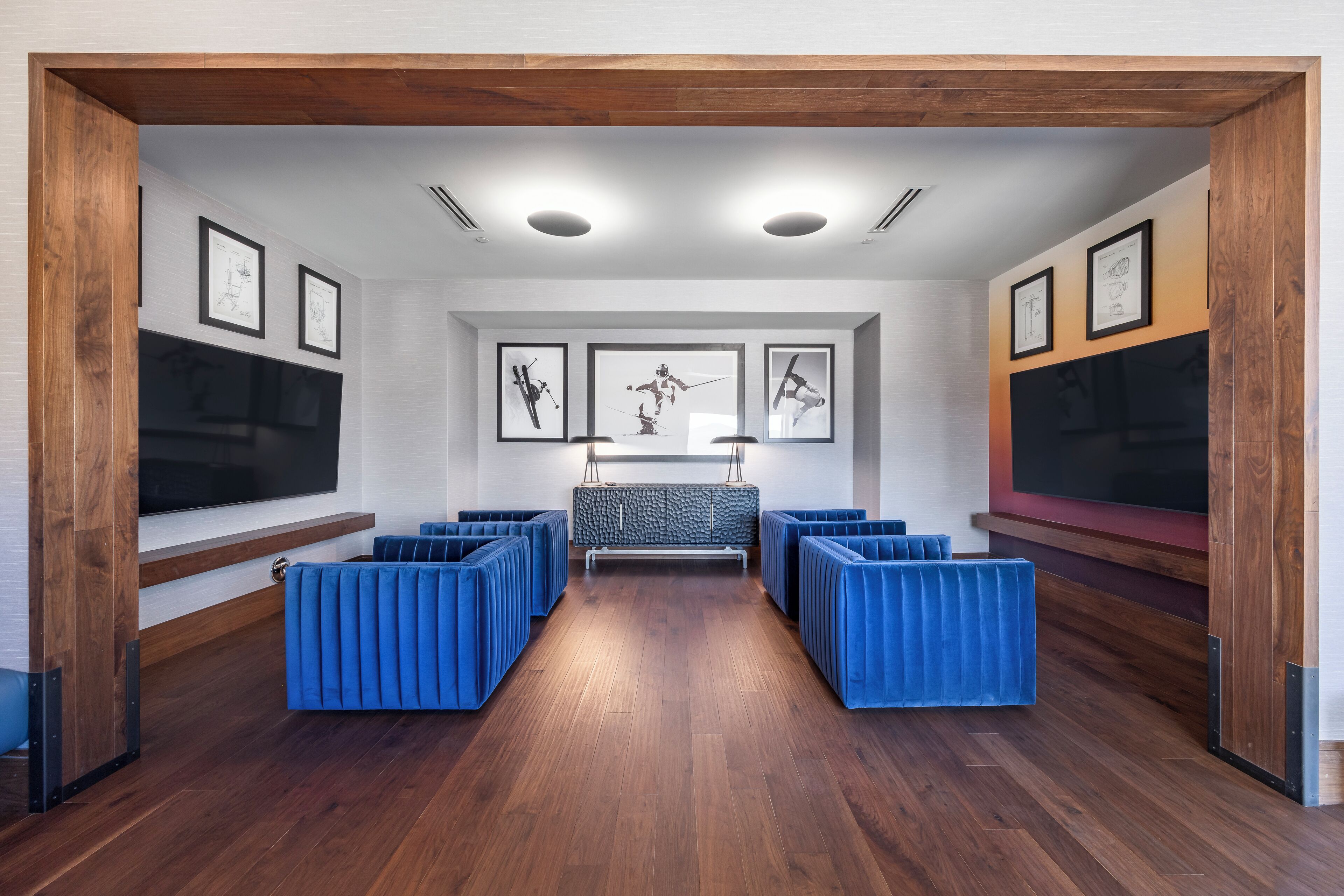 Quarto moderno com piso de madeira, assentos azuis, uma televisão grande e obras de arte emolduradas nas paredes.