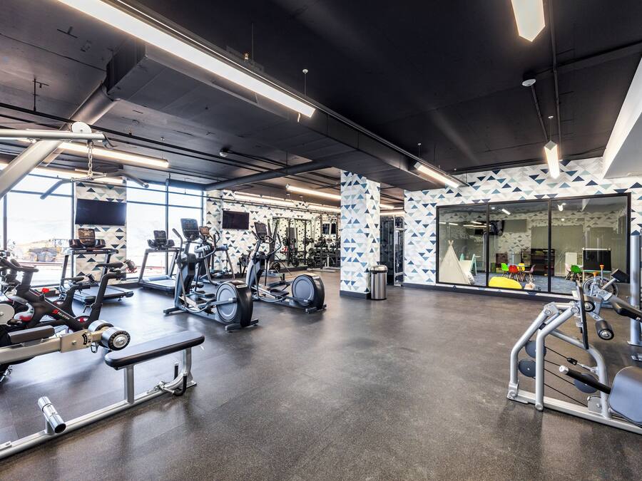 A modern gym with various exercise equipment, large windows, and a patterned wall.
