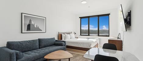 Una habitación moderna y luminosa con un sofá gris, una mesa de centro de madera, una cama con ropa de cama blanca y un gran ventanal con vistas al cielo y al paisaje distante.