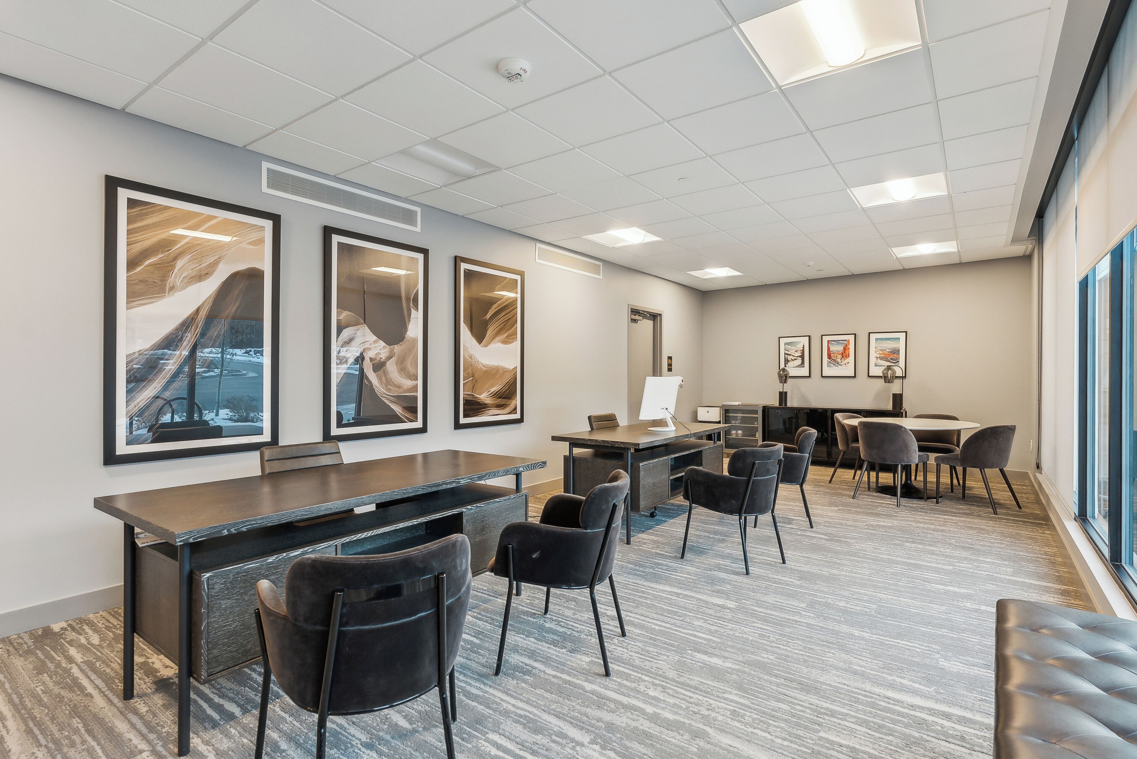 A modern office space with a long desk, several chairs, framed artwork on the wall, and a large window.