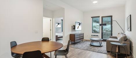 Una sala de estar moderna con una mesa redonda de madera, sillas negras, un sofá gris y un televisor sobre un mueble de madera.