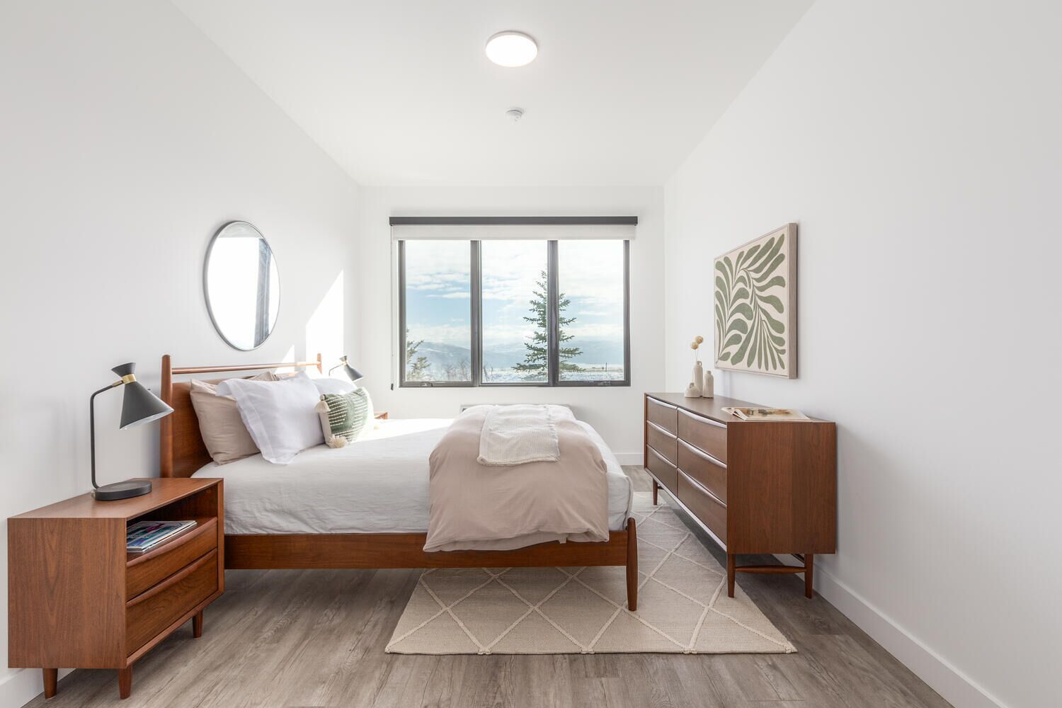 A modern bedroom with a wooden bed, a bedside table, a dresser, a large window with a view, and a ceiling light.