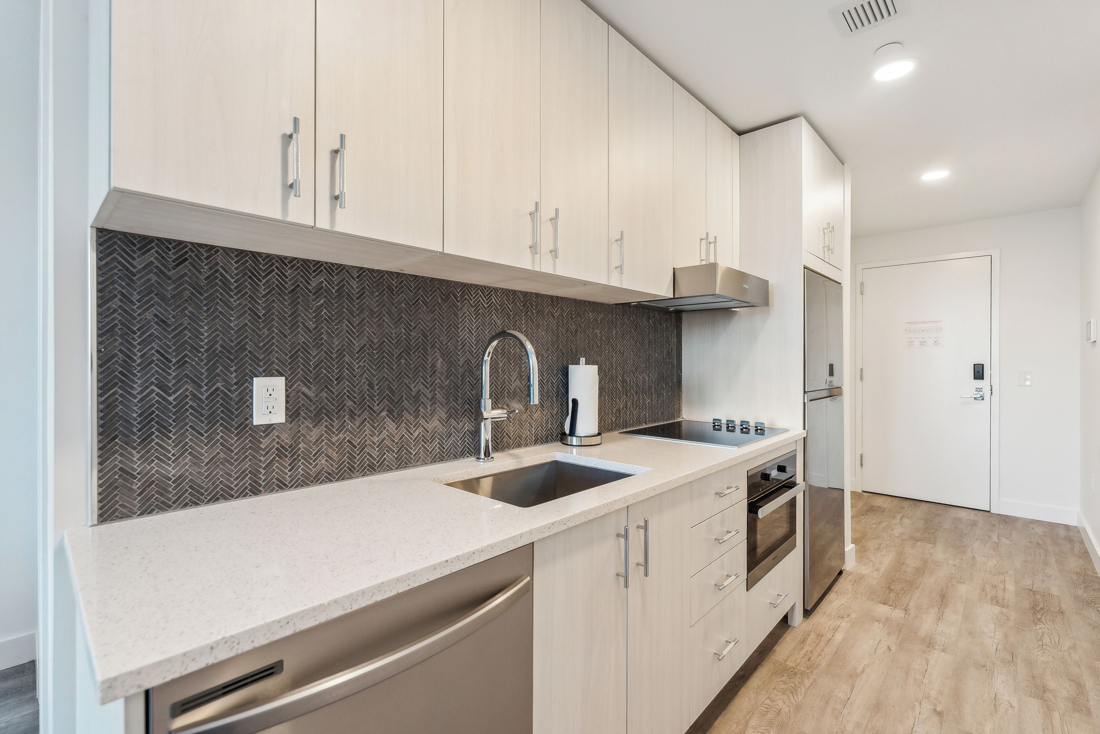 A modern kitchen with a herringbone-patterned backsplash, stainless steel appliances, and light wood cabinetry.