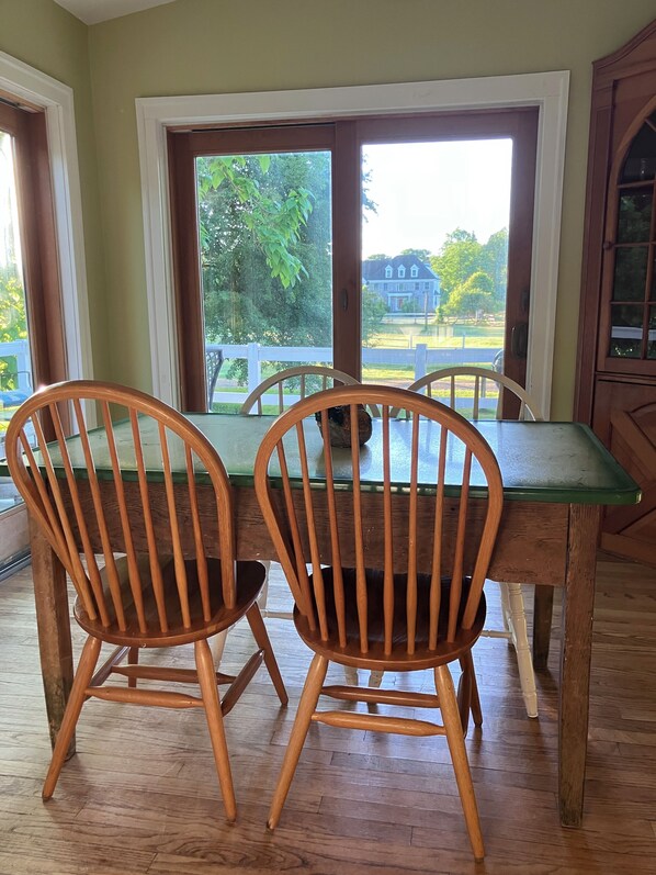 A dining area with a wooden table and chairs, large windows with a view of a house and greenery.