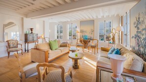 A spacious living room with a high ceiling, wooden flooring, and large windows allowing natural light. There are multiple seating areas with sofas and chairs, and a dining table with chairs. - 2 storey luxury penthouse with large terrace but no elevator (Paris)