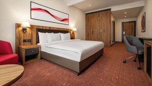 A hotel room with a large bed, two bedside tables with lamps, a red armchair, a desk with a chair, and a wooden wardrobe.