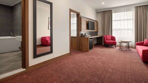A hotel room with a large mirror, a red sofa, a small round table, a TV mounted on the wall, and a bathroom with a bathtub.